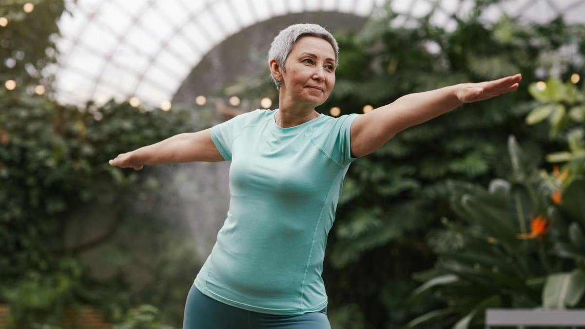 Elderly woman performing a yoga warrior pose in a lush green indoor botanical garden.