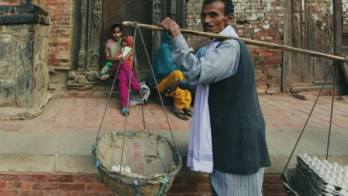 An Indian man carries a traditional balance scale in a village street, with cultural architecture in the background.