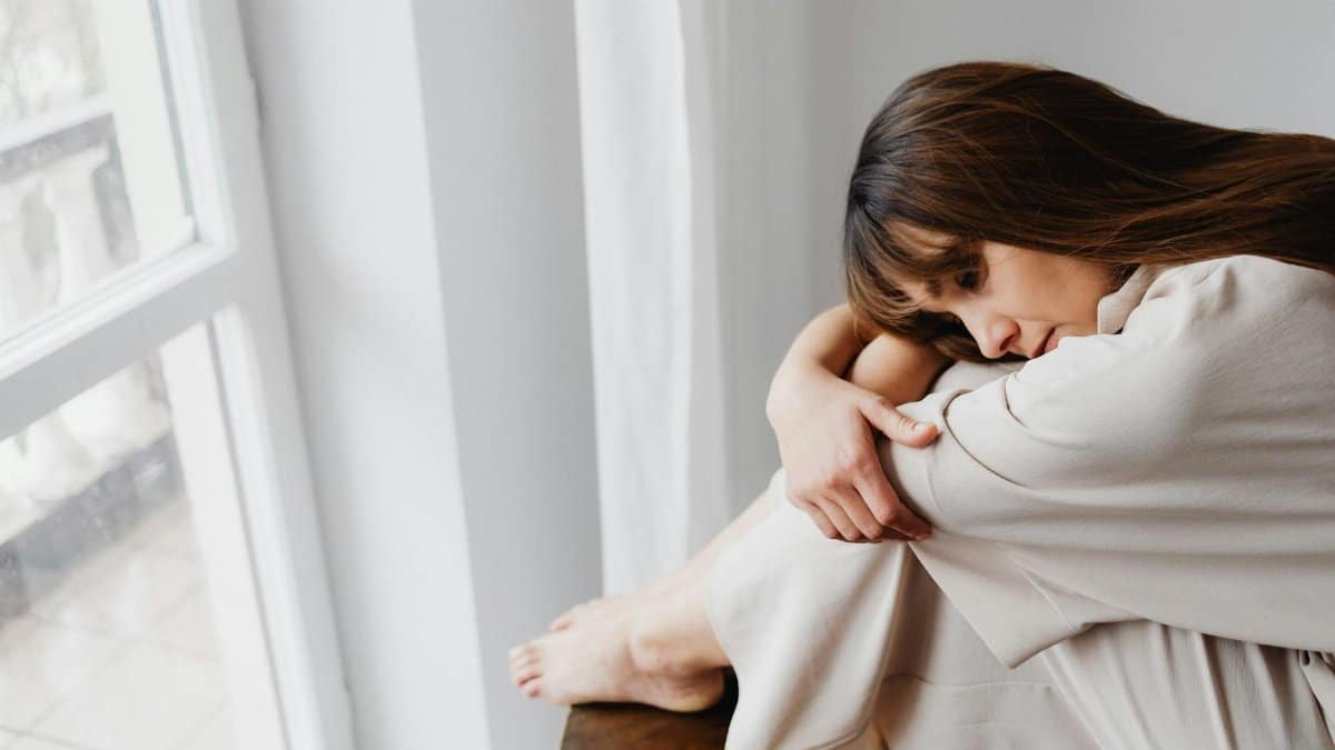 Thoughtful woman in neutral attire sitting by a window, reflecting emotions.