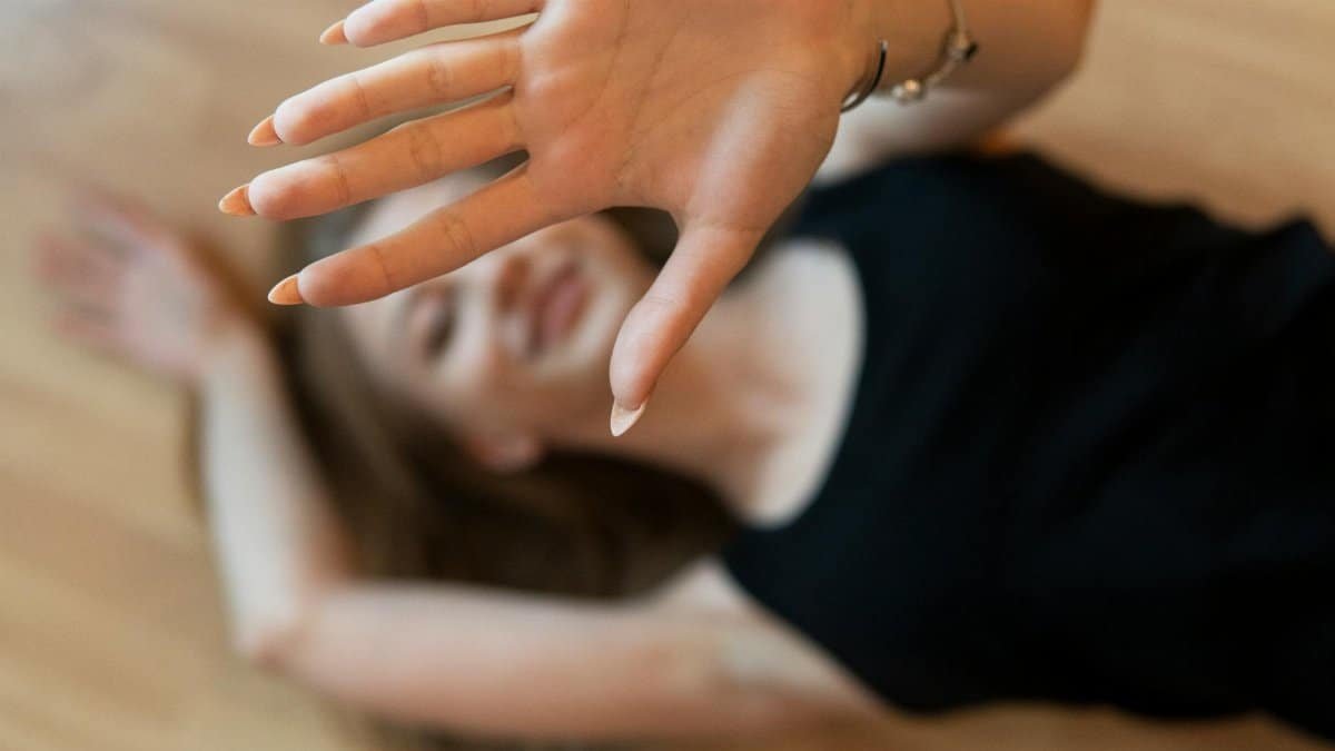 A woman lying on a wooden floor with a hand gesture blocking the frame, smiling naturally.
