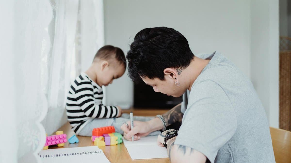 Father writing while son plays with toys at the table in a cozy room.
