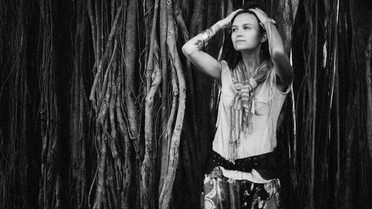 A woman stands against dramatic tree roots, hands on head, in grayscale.