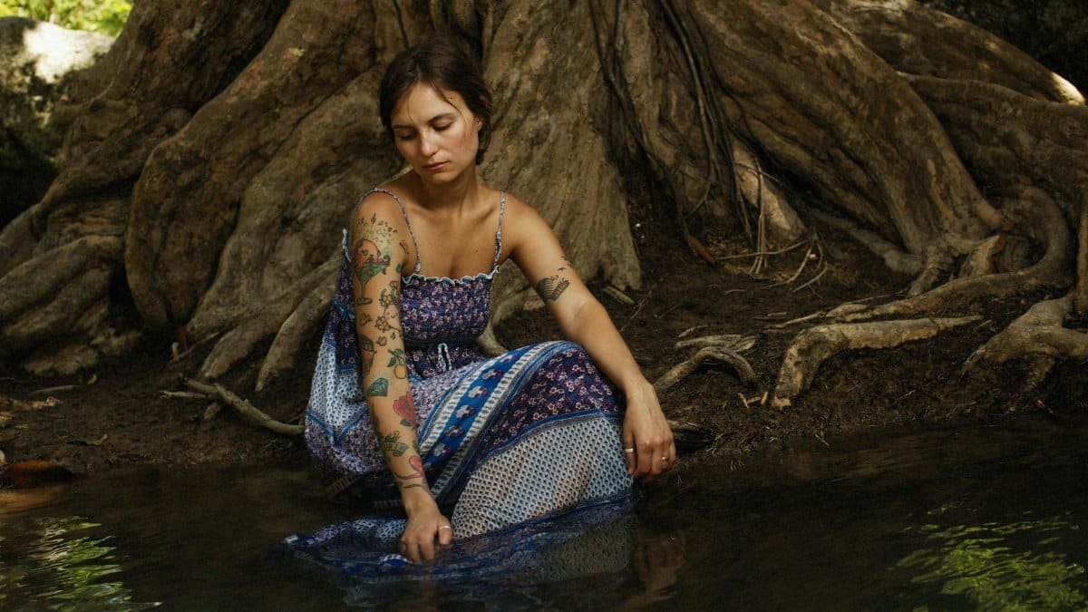 A young tattooed woman sits pensively by a stream among tree roots, reflecting.