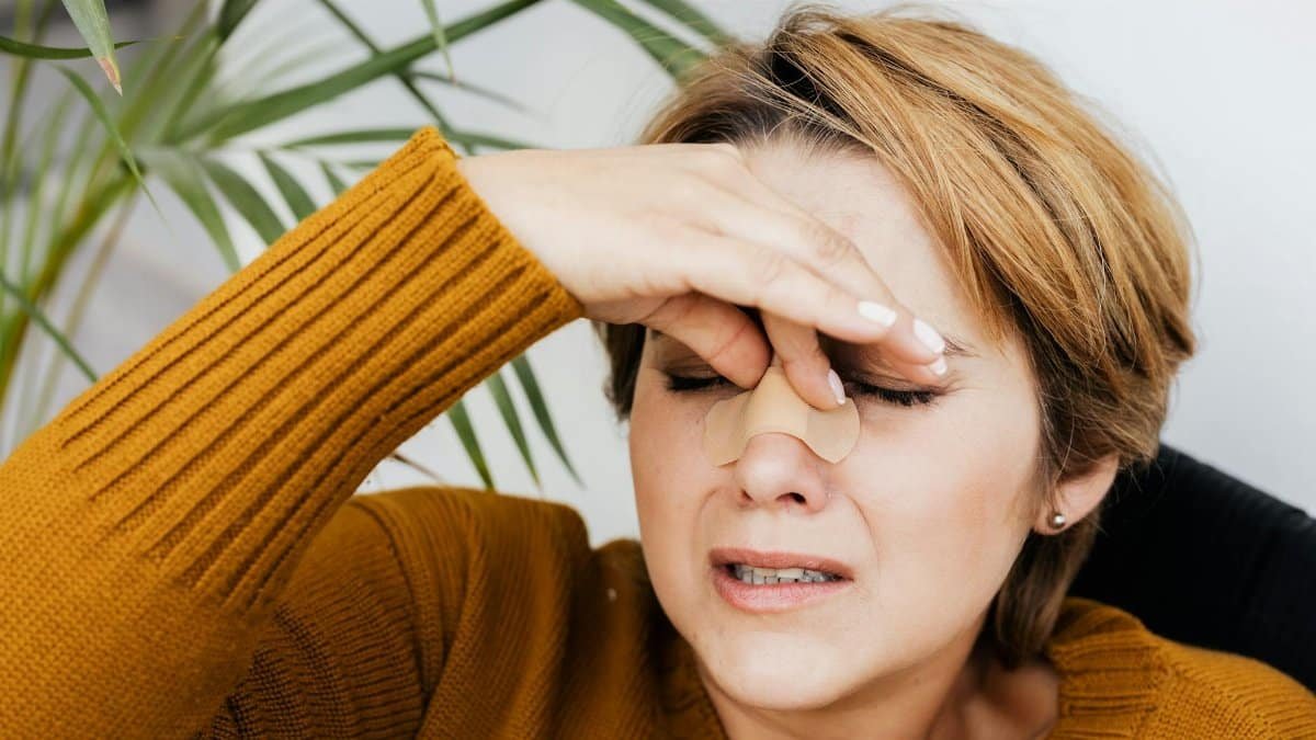 Woman holding her nose with a band-aid, showing discomfort and pain indoors.