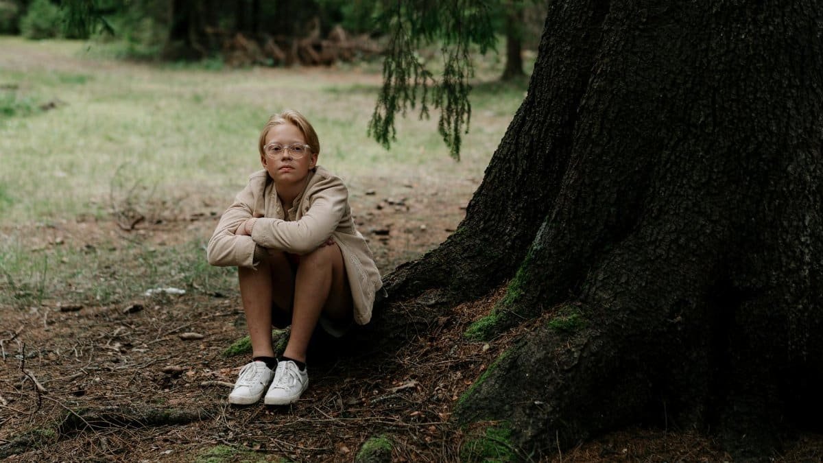 A teenager sits pensively by a large tree in a quiet forest area.