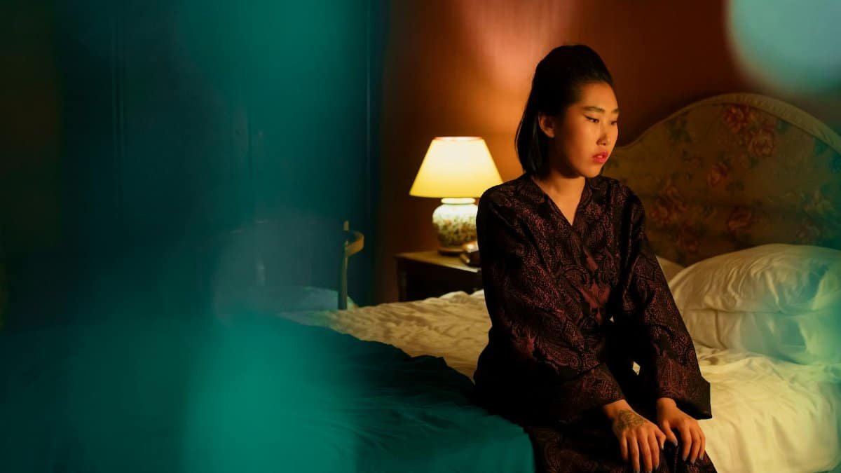 A woman in a robe sits thoughtfully on a bed in a warmly lit elegant bedroom.