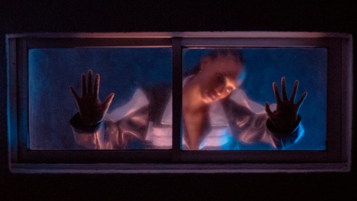 A woman places her hands on a foggy window, illuminated by dramatic lighting.