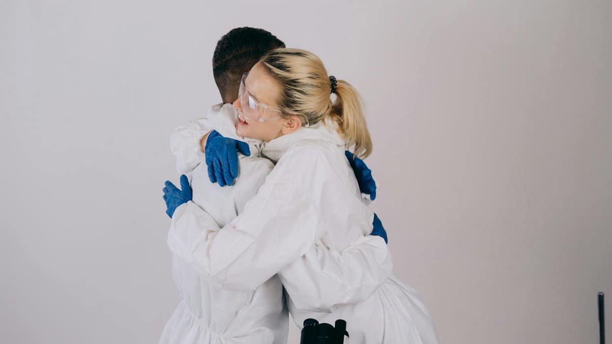 Two scientists hugging while wearing protective gear and goggles in a lab setting.