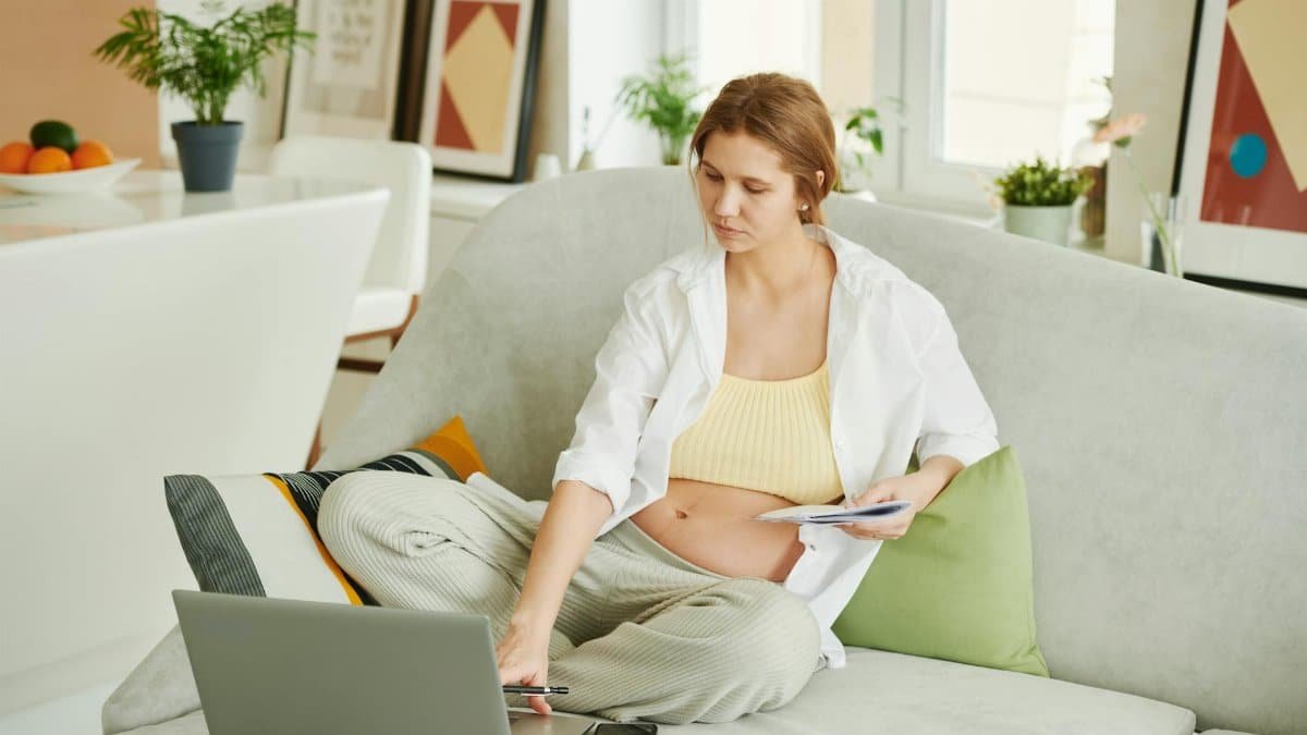 Pregnant woman sitting on sofa working on laptop in cozy home setting.