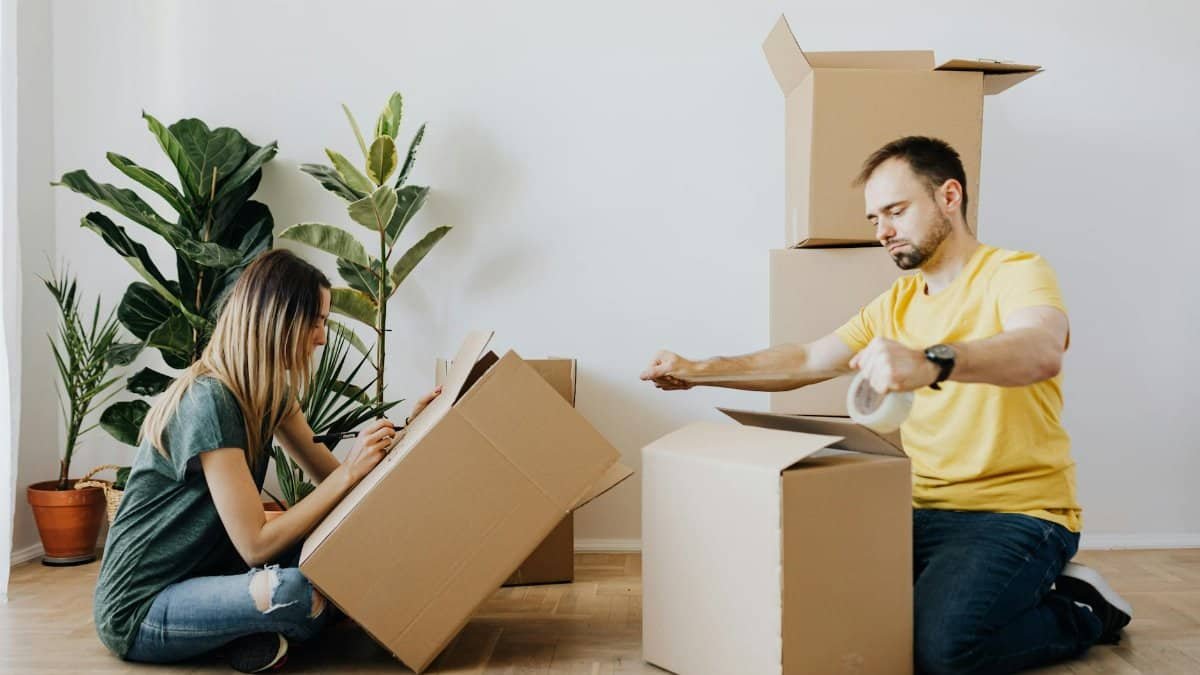 Side view full body unemotional couple wearing casual clothes and sitting on parquet while sealing carton boxes with tape and writing notes during moving in together