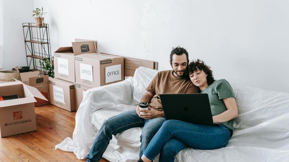 A couple sits together on a couch, using a laptop amidst moving boxes, enjoying relaxed time indoors.