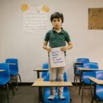 Child standing on desk in classroom holding a sign against bullying, promoting kindness and awareness.