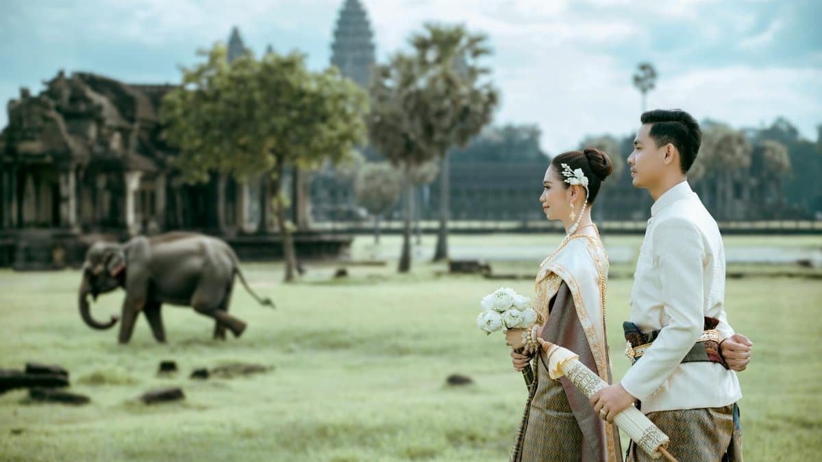 Elegant Cambodian couple in traditional attire at Angkor Wat with an elephant in the background.