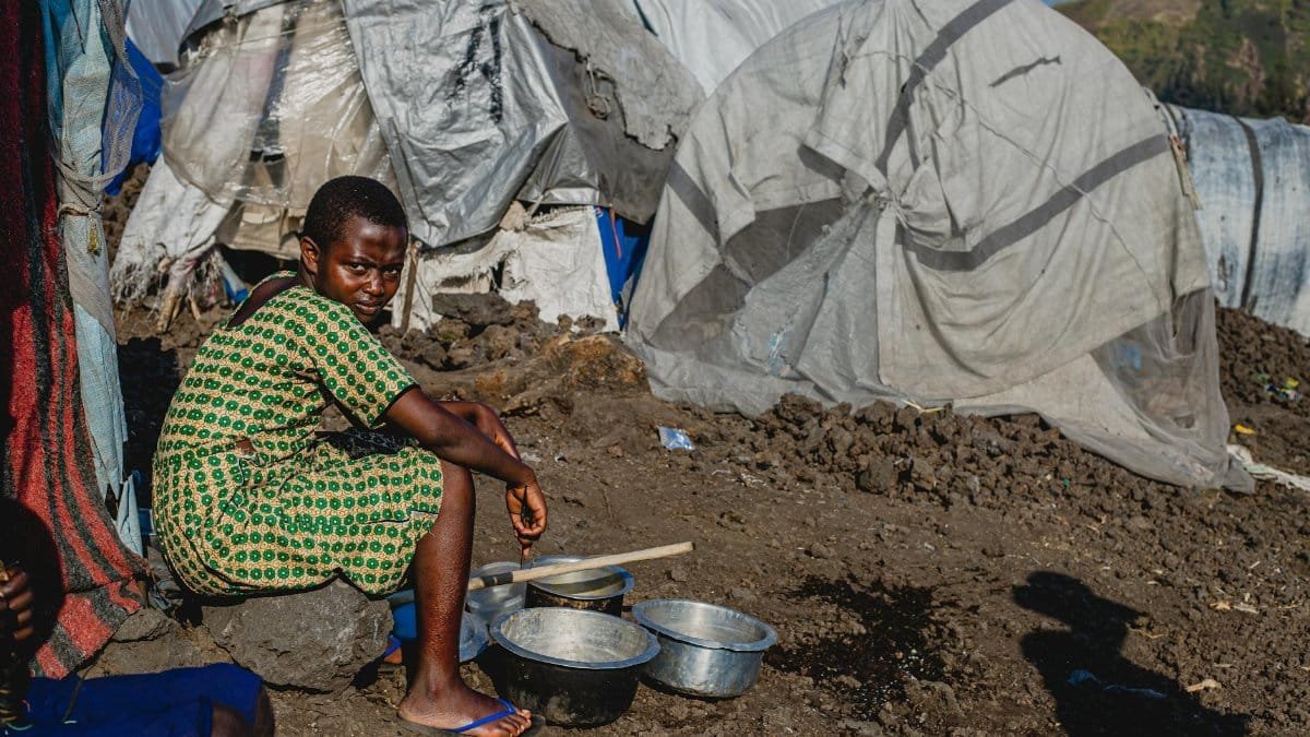 A young girl sits outside a makeshift shelter with metal bowls, highlighting resilience.