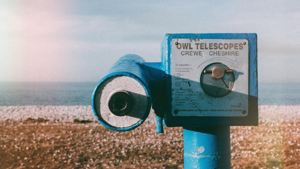 Vintage blue telescope by a pebble beach with ocean view under a clear sky.