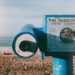 Vintage blue telescope by a pebble beach with ocean view under a clear sky.