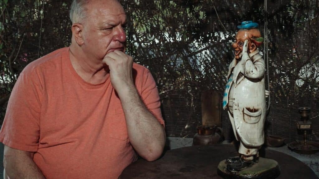 An elderly man in deep thought sits next to a whimsical statue outdoors.