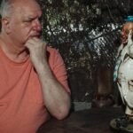 An elderly man in deep thought sits next to a whimsical statue outdoors.