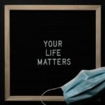 Overhead arrangement of framed black photo with white words Your Life Matters on black background near medical mask