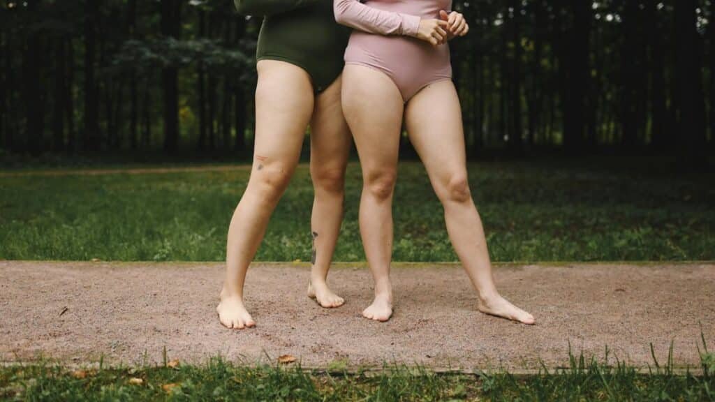 Two women in bodysuits embracing body positivity outdoors on grass.