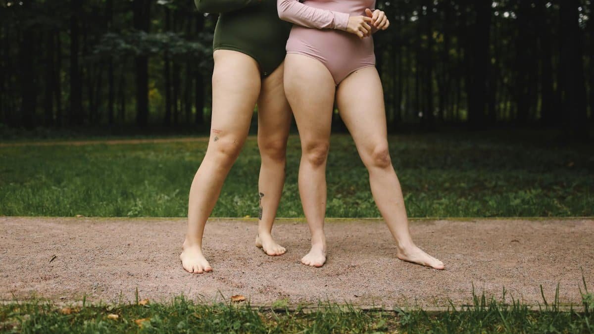 Two women in bodysuits embracing body positivity outdoors on grass.