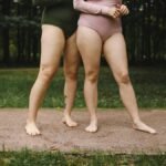 Two women in bodysuits embracing body positivity outdoors on grass.