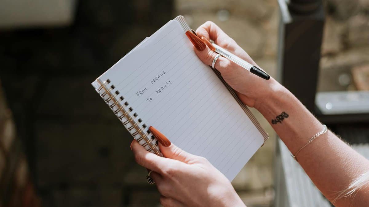 A woman with tattooed arm writes 'From dreams to reality' in a notebook.