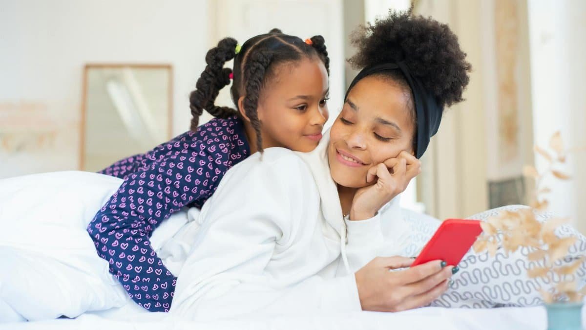 A heartwarming moment between a mother and daughter embracing while using a smartphone indoors.