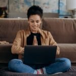 A smiling young woman sits cross-legged on a pouf, using her laptop in a cozy home setting.