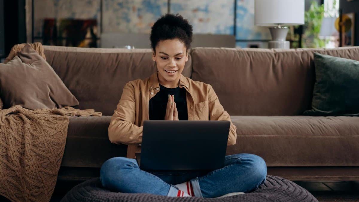 A smiling young woman sits cross-legged on a pouf, using her laptop in a cozy home setting.