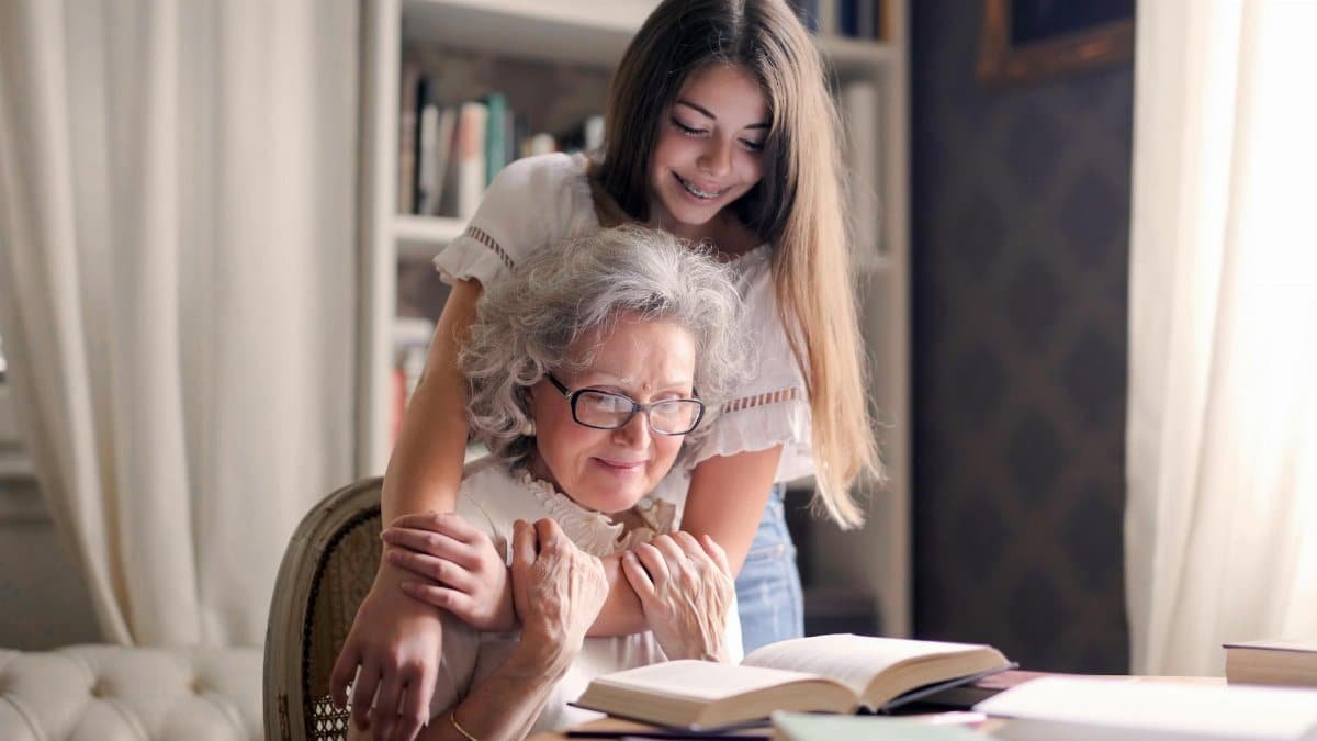A heartwarming moment of a granddaughter embracing her grandmother as she reads a book together indoors.