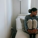 A young woman sits alone on a bed, looking upset and lonely in a cozy room.