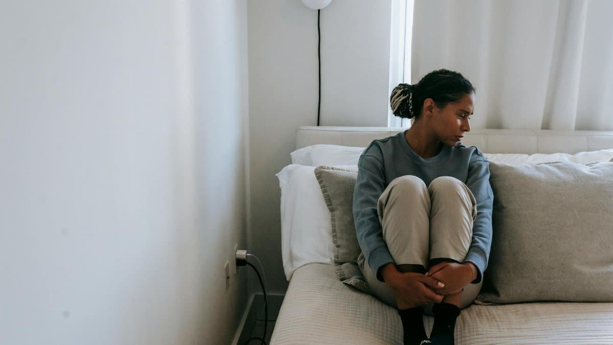 A young woman sits alone on a bed, looking upset and lonely in a cozy room.