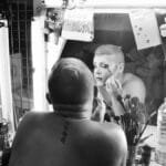 Drag queen applying makeup in dressing room, showcasing artistry and transformation.