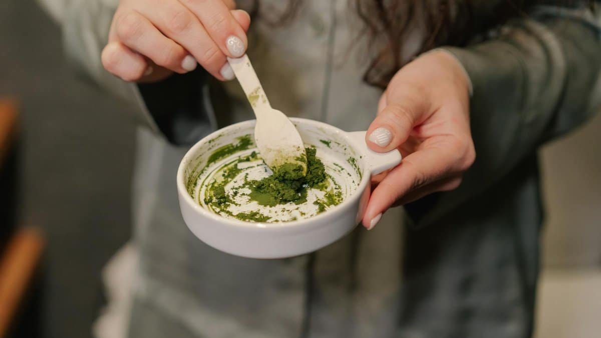 Close-up of a woman preparing a green paste in a ceramic bowl, focusing on hands and texture.