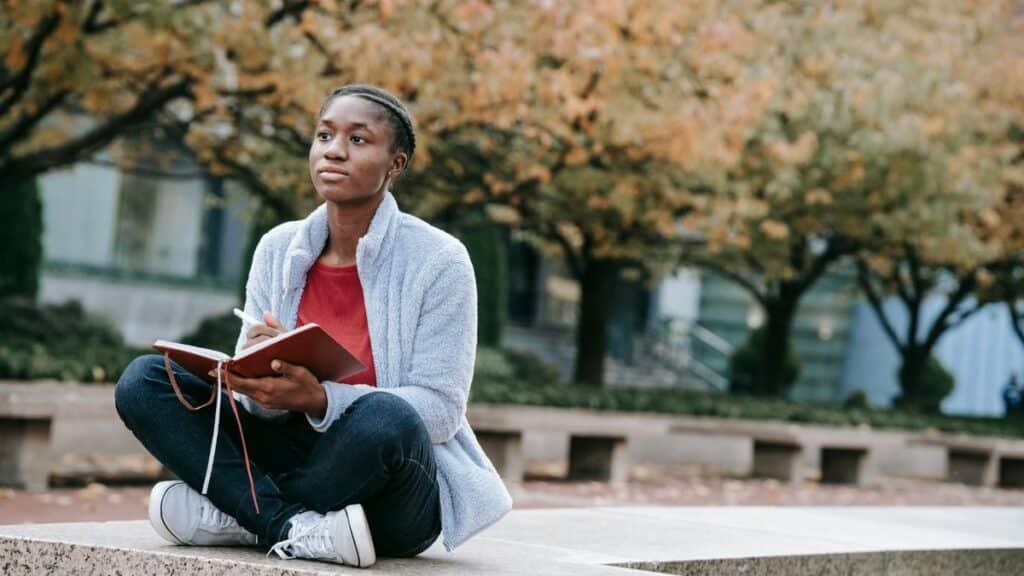 Young contemplative African American woman with agenda sitting with crossed legs against autumn trees while looking forward in city