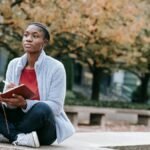 Young contemplative African American woman with agenda sitting with crossed legs against autumn trees while looking forward in city