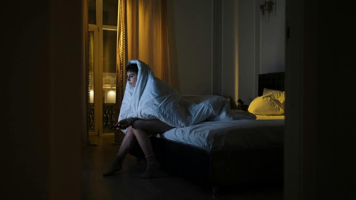Woman wrapped in blanket watching TV, battling insomnia in dimly lit bedroom.