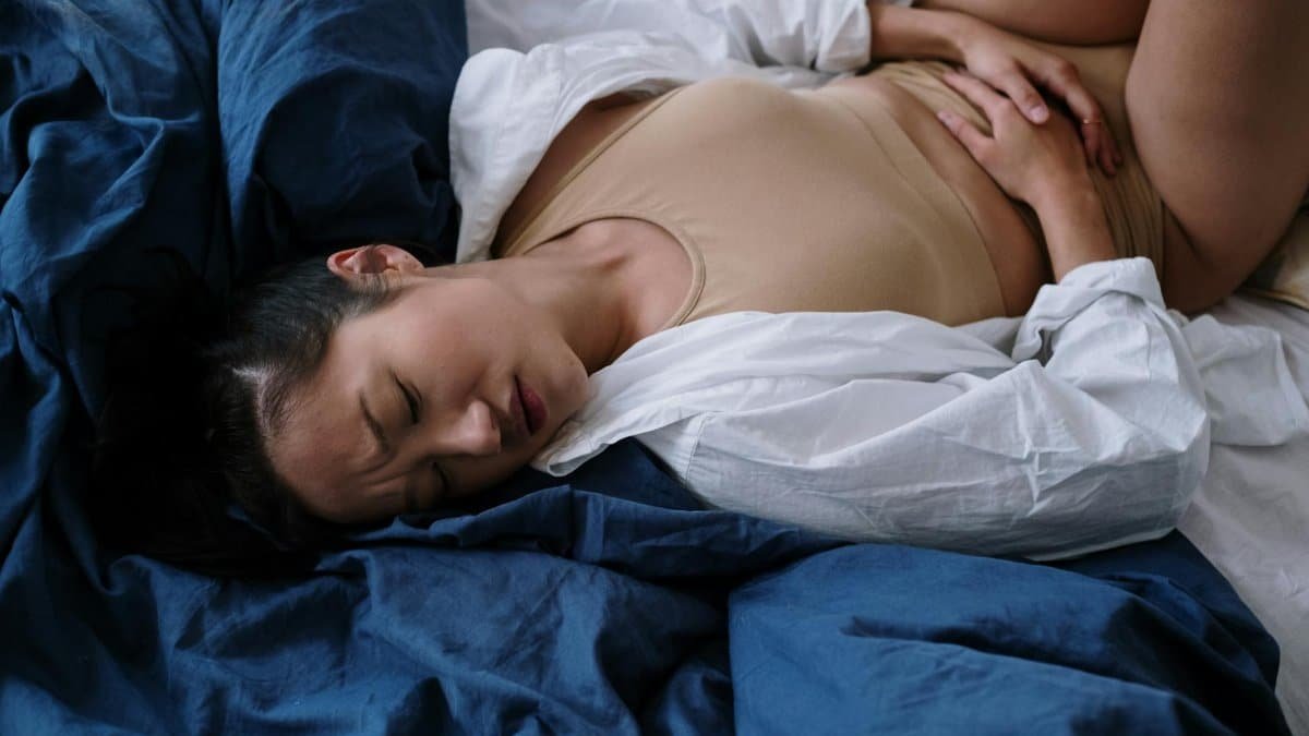 A woman lying on a bed in discomfort due to stomach pain, expressing symptoms of illness.