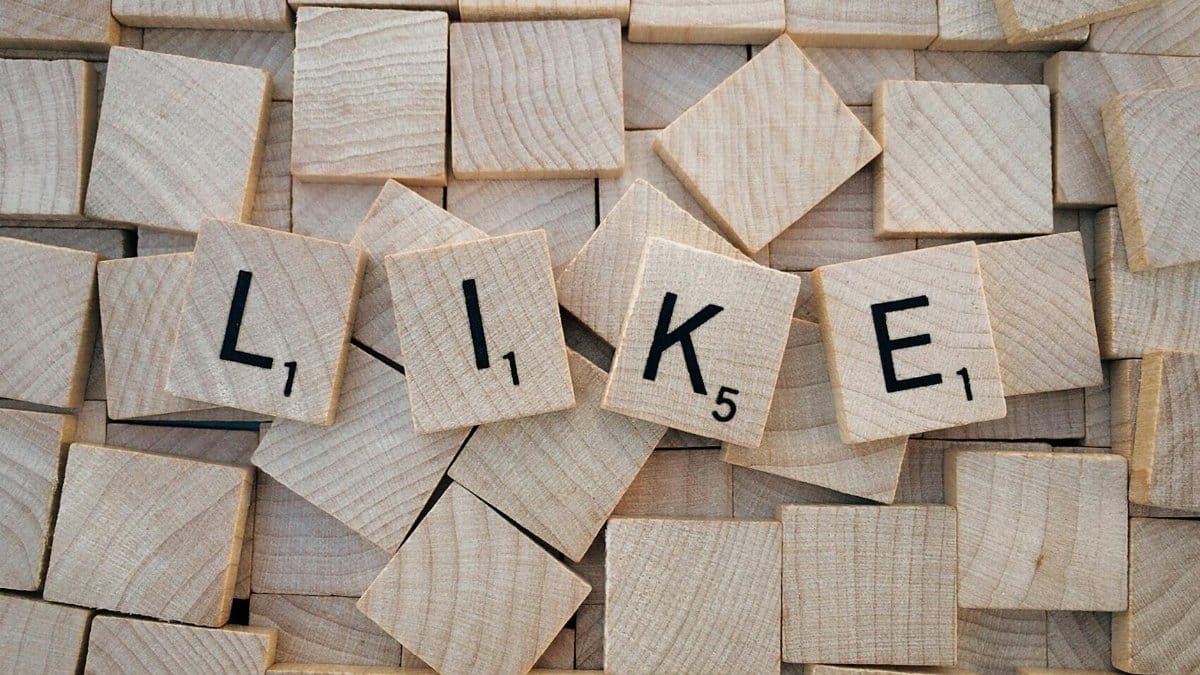 Top-down view of Scrabble tiles spelling 'LIKE' on a pile of blank wooden tiles.
