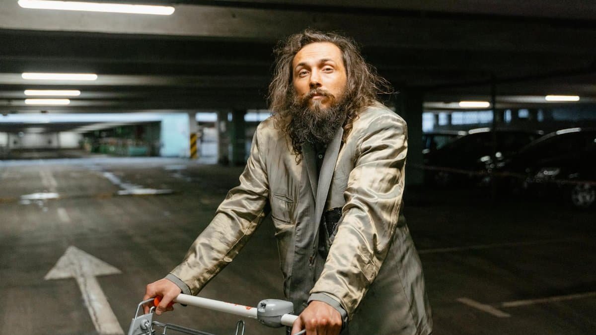 A homeless man with a beard and worn clothes pushes a shopping cart in a dimly lit parking garage.