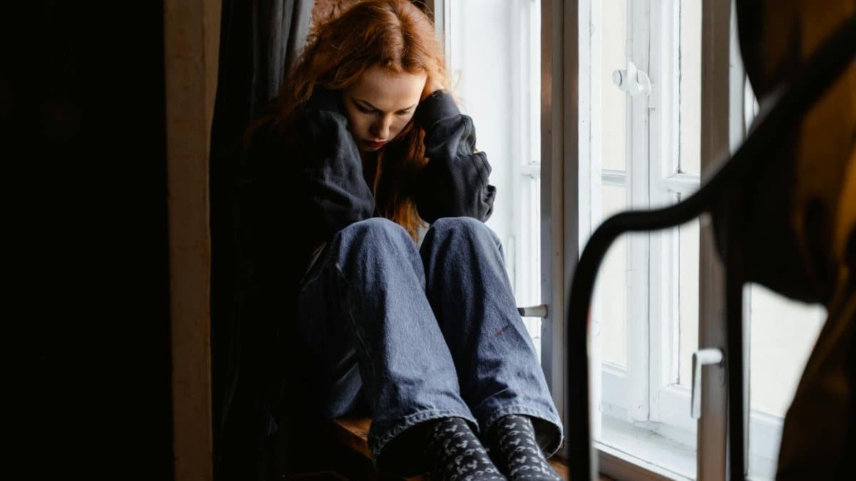 A woman sits pensively by a window, expressing feelings of solitude and contemplation.