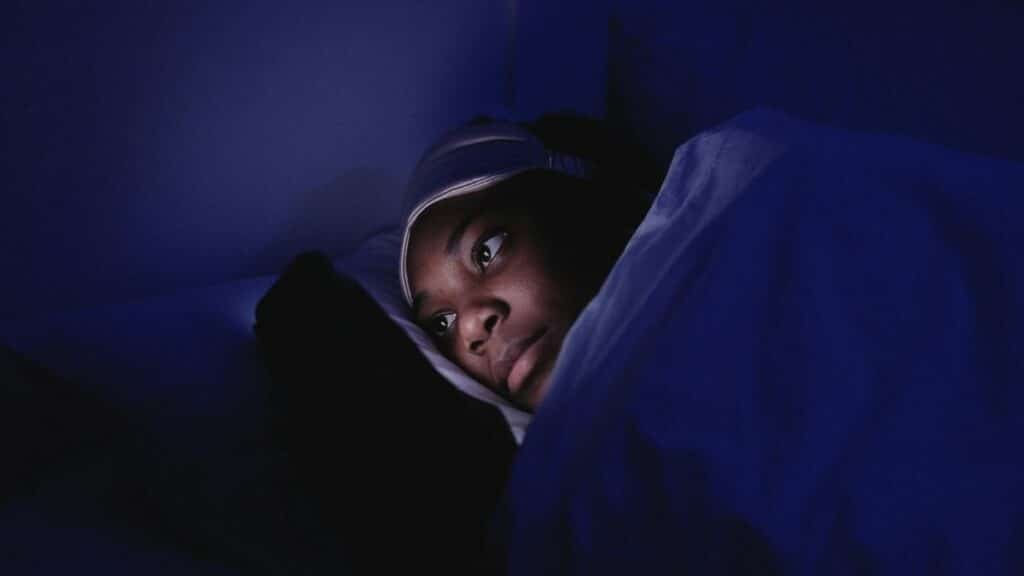 A woman lies in bed at night using her smartphone, illuminated by its screen.