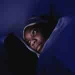 A woman lies in bed at night using her smartphone, illuminated by its screen.