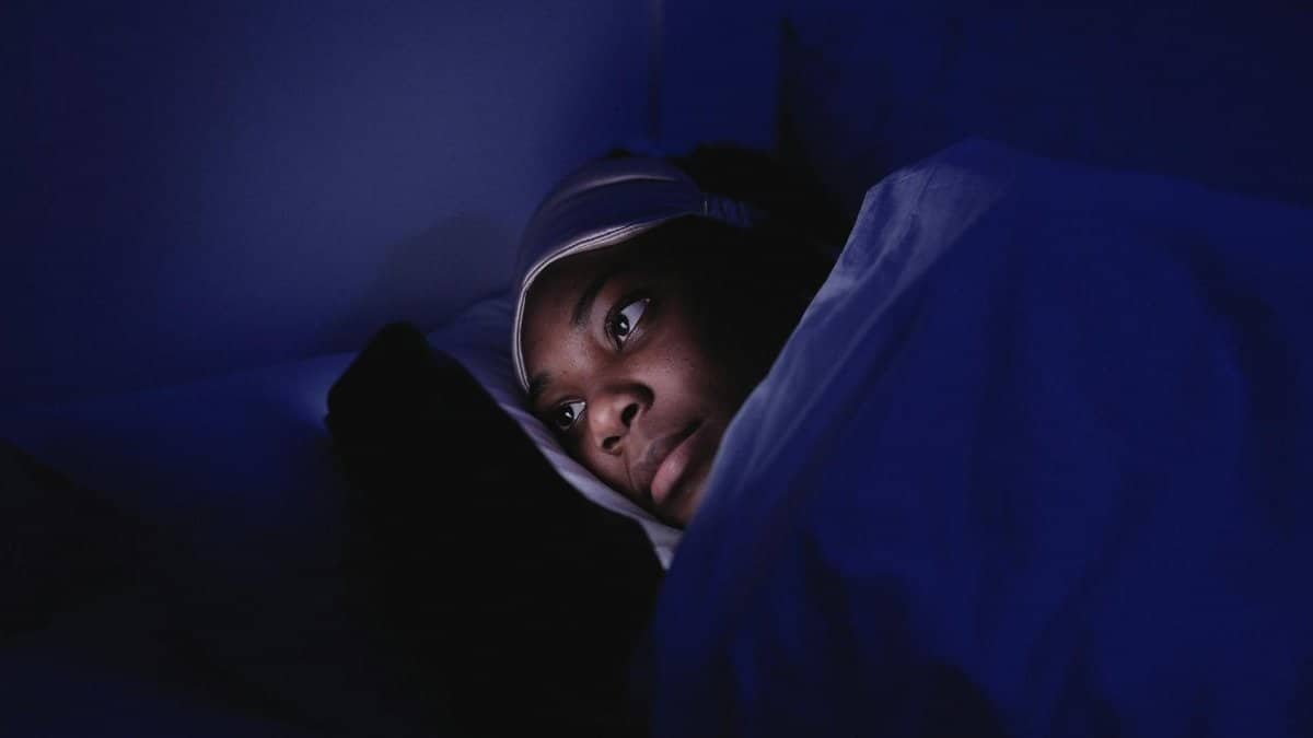 A woman lies in bed at night using her smartphone, illuminated by its screen.