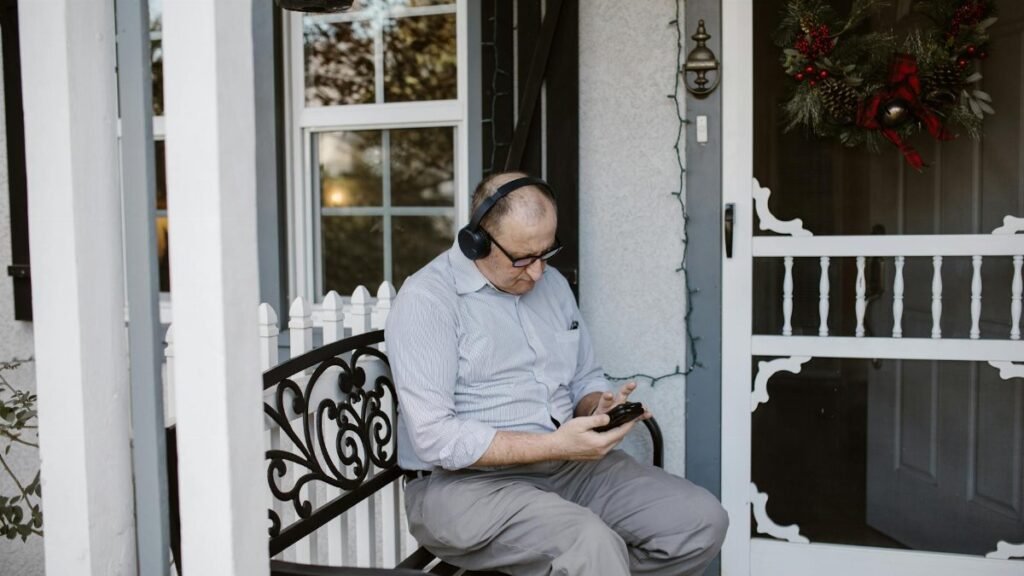 Elderly man enjoying music on porch, wearing headphones and checking phone.