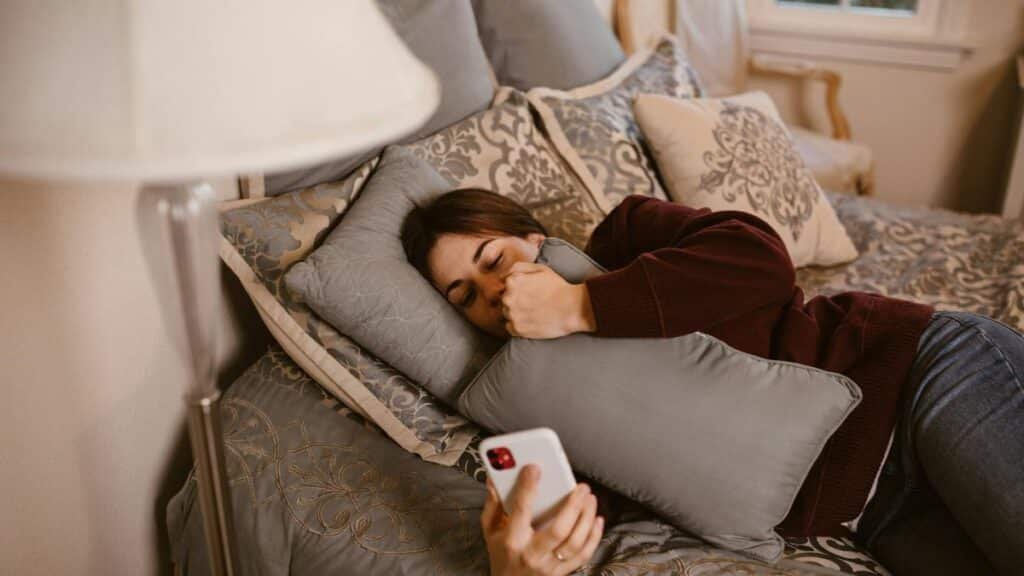 A young woman lying on a bed, holding a phone and a pillow, symbolizing relaxation and connection.