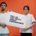 Two people holding a 'Tell Us The Truth About Coronavirus' sign against a vibrant orange background.