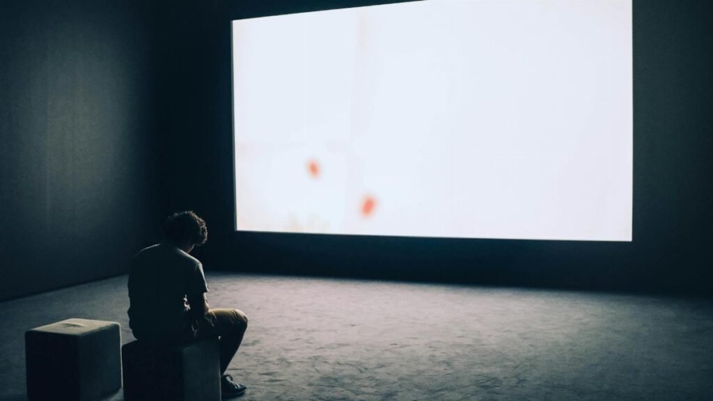 A man sits alone in a dark room with a large white screen, depicting solitude and contemplation.