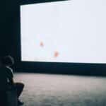 A man sits alone in a dark room with a large white screen, depicting solitude and contemplation.